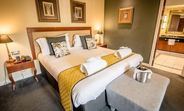 Hotel bedroom at Wild Pheasant Hotel & Spa featuring a neatly made bed, towels folded on the bed, slippers, a table with champagne.